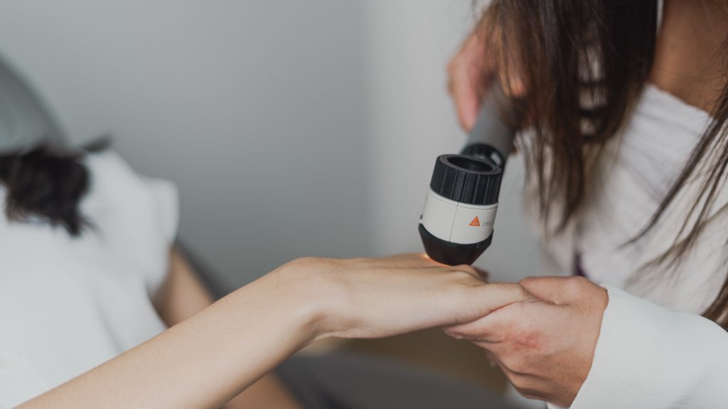 Mão da dermatologista apoia o dermatoscópio no punho de uma paciente durante o exame