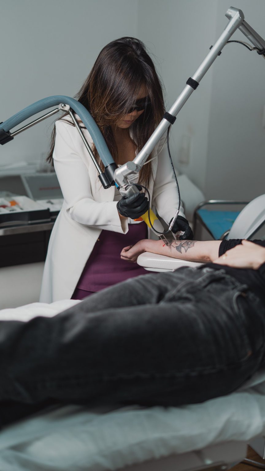 Dra. Juliana Toma, de óculos de proteção, conduz o laser de picossegundos em uma paciente deitada, na sala de laser da clínica
