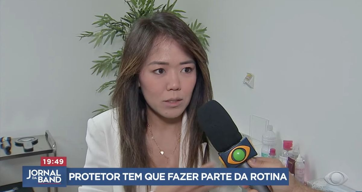 Dra. Juliana Toma entrevistada pelo Jornal da Band no consultório, com o microfone da Band e a legenda “Protetor tem que fazer parte da rotina”